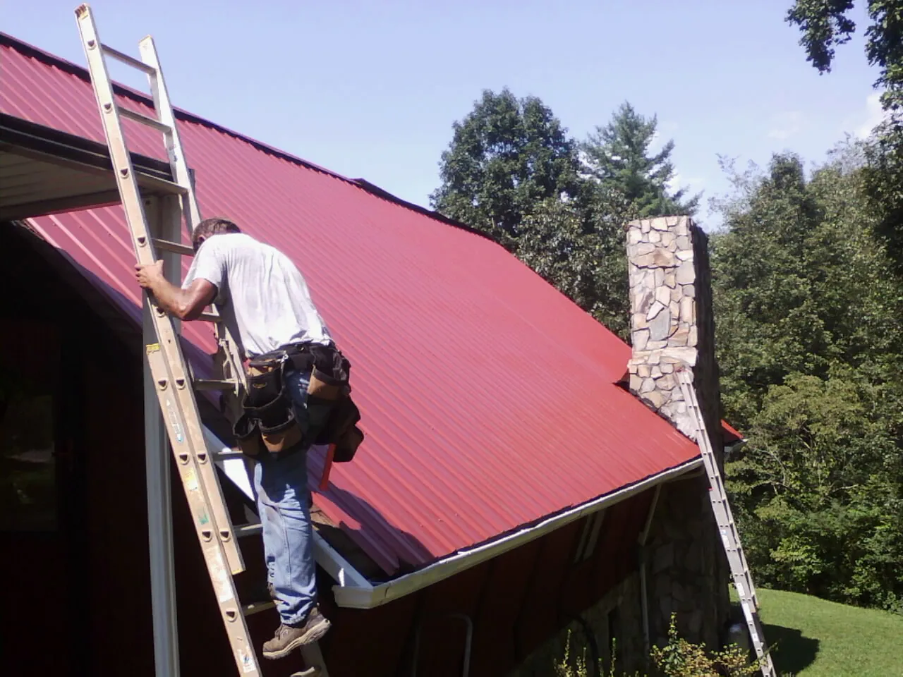 Roofer on a ladder beside a red metal roof and stone chimney — Soffit & Fascia Repair by Roofing Brothers in Inkster